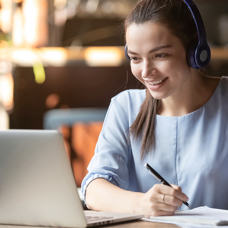 woman in front of laptop with headphones taking notes while smiling